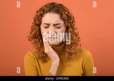 Young and attractive caucasian redhead girl touching her mouth with toothache isolated on orange studio background. Stock Photo