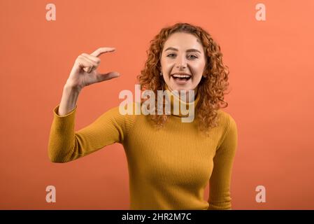 Young and attractive redhead Caucasian girl in orange jumper showing small size with her fingers isolated on orange studio background. Stock Photo