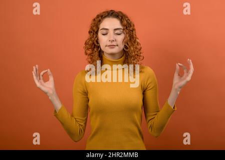 Young and attractive redhead Caucasian girl in orange jumper holding hands in mudra gesture isolated on orange studio background. Meditation, yoga, we Stock Photo