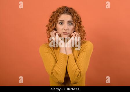 Portrait of a frightened and shocked redhead Caucasian girl in orange jumper isolated on orange studio background. Stock Photo