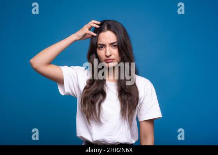 Attractive caucasian or arab brunette girl in a white t-shirt holding her hand on her head and thinking isolated in on a blue studio background. Stock Photo