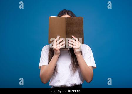 Attractive brunette girl in a white t-shirt holding a book and reading isolated on a blue studio background. Concept of knowledge and learning. Stock Photo