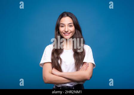 Portrait of happy and attractive caucasian or arab brunette girl in white t-shirt with crossed arms isolated on blue studio background. Stock Photo