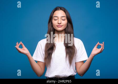 Attractive caucasian or arab brunette girl in white t-shirt meditating in lotus position and showing zen gesture isolated on blue studio background. Stock Photo