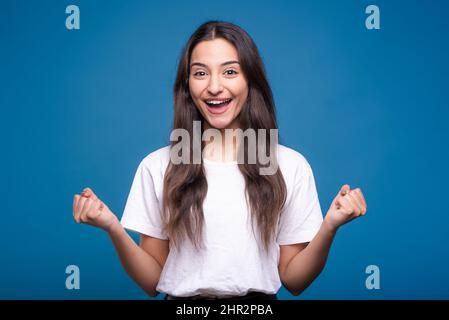 Attractive caucasian or arab brunette girl in white t-shirt showing winner gesture clenching fists isolated on blue studio background. Stock Photo