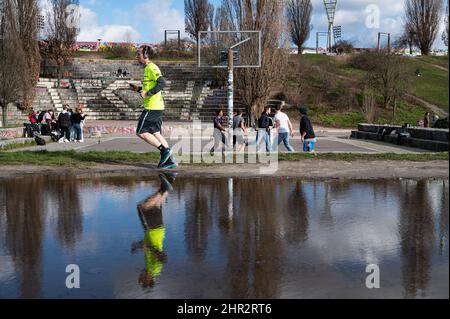 23.02.2022, Berlin, Germany, Europe - Man is reflected in puddle as he jogs past a group of young men playing basketball in the backdrop at Mauerpark. Stock Photo