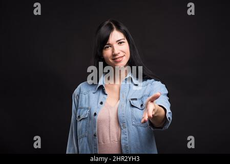 Attractive caucasian brunette girl in shirt showing hand gesture greeting, handshake isolated on black studio background. Stock Photo