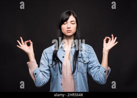 Attractive caucasian brunette girl in shirt in lotus position showing zen and mudra gesture isolated on black studio background. Stock Photo