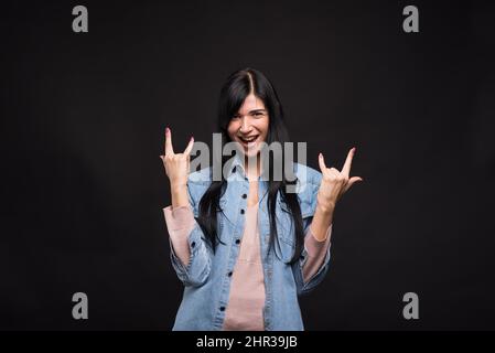 Attractive caucasian brunette girl in shirt happily screaming and showing rocker or punk gesture isolated on black studio background. Stock Photo