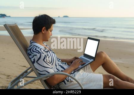 Lifestyle of young Asian man blogger working and using keyboard on laptop while sitting on the beautiful beach, freelance travel on holiday summer dur Stock Photo