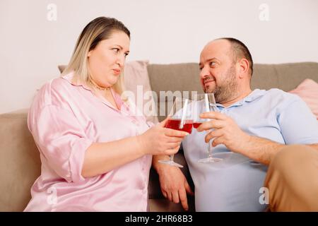 Happy caucasian couple relax on cozy sofa at home resting holding in their hands two glasses of wine and looking at each other with love. Stock Photo