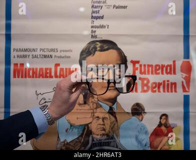 Bonhams, London, UK. 25 February 2022. Sir Michael Caine: The Personal Collection preview before sale on 2 March 2022. He and his wife, Lady Caine, are downsizing and have put some of their treasured possessions up for auction. Image: Funeral In Berlin, signed film poster, Paramount, 1966, estimate: £200-300 and a pair of the actors iconic spectacles. Credit: Malcolm Park/Alamy Live News Stock Photo
