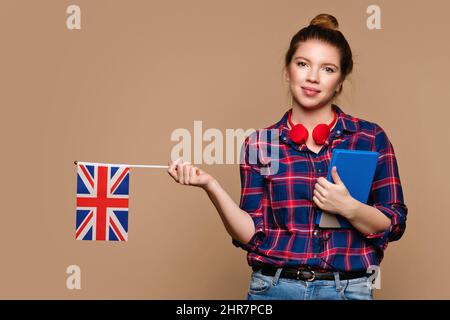 Smart Student girl holds small UK flag and notebook in studio on beige background. Study abroad concept. International student exchange program. Learning British English with native speaker. Stock Photo