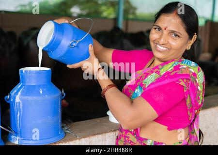 Happy smiling Indian woman busy working by pouring milk into container while looking at camera - concept of milk production, agri business, growth and Stock Photo