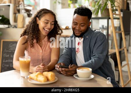 Happy african american young couple sharing smartphone while sitting at coffee shop Stock Photo