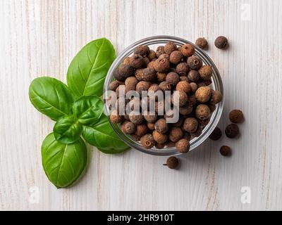 Glass bowl full of allspice jamaica pepper near fresh basil leaves on a white wooden table. Dried berries of the Pimenta dioica tree for cooking. Stock Photo