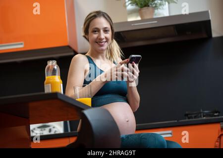 Pregnant blonde woman drinking an orange juice in the kitchen and using phone Stock Photo