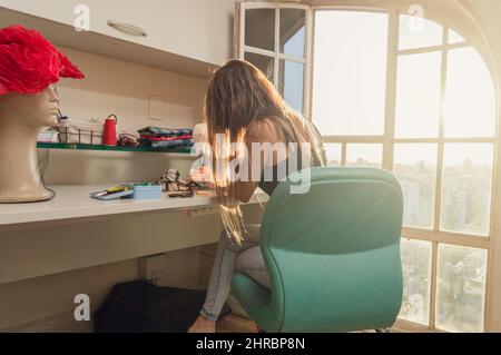 young latin caucasian woman by her window using her sewing machine in her sewing studio, sewing fabrics designing and the sunlight enters through the Stock Photo