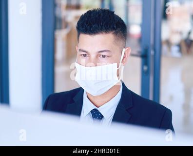 First rule of office work Wear a face mask. Shot of a young businessman wearing a face mask and using a computer in a modern office. Stock Photo
