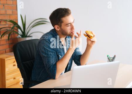 Hungry freelance programmer male eating hamburger with beef from fast food restaurant watching online cinema on laptop computer Stock Photo