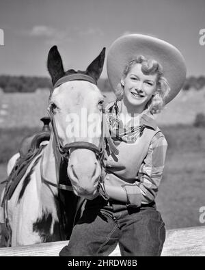 1940s SMILING BLONDE COWGIRL WEARING COWBOY HAT BLUE JEANS SITTING ON FENCE HOLDING ONTO HER HORSE’S HALTER LOOKING AT CAMERA - h1158 HAR001 HARS WEST COTTON PLEASED JOY LIFESTYLE FEMALES RURAL HOME LIFE COPY SPACE HALF-LENGTH LADIES PERSONS CHARACTER WESTERN PLAID DENIM VEST B&W EYE CONTACT COWBOYS HAPPINESS MAMMALS CHEERFUL ADVENTURE RECREATION COWGIRL HALTER OCCUPATIONS ONTO SMILES COWGIRLS JOYFUL STYLISH BLUE JEANS HORSE'S INFORMAL MAMMAL TWILL YOUNG ADULT WOMAN BANDANNA BLACK AND WHITE CASUAL CAUCASIAN ETHNICITY HAR001 OLD FASHIONED Stock Photo