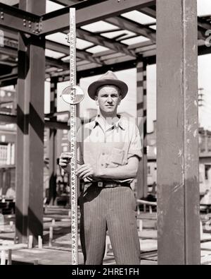 1940s MAN ON CONSTRUCTION SITE HOLDING A LEVELING OR GRADING ROD MEASURED IN DECIMAL FEET FOR USE IN SURVEYING - i43 HAR001 HARS COOPERATION MID-ADULT MID-ADULT MAN PRECISION BLACK AND WHITE CAUCASIAN ETHNICITY HAR001 LABORING OLD FASHIONED Stock Photo