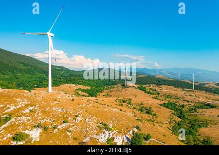 Windmills with large propellers produce green energy in highland against forestry mountains under blue sky on sunny summer day aerial view Stock Photo