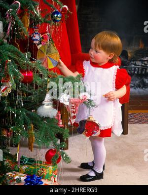 1980s LITTLE GIRL WEARING RED DRESS WHITE PINAFORE MARY JANE PATENT LEATHER SHOES FOCUSED ON ORNAMENT ON CHRISTMAS TREE  - kx8839 PHT001 HARS JUVENILE CUTE STYLE MYSTERY DECORATIONS JOY LIFESTYLE CELEBRATION FEMALES HOME LIFE COPY SPACE FULL-LENGTH INSPIRATION ORNAMENT SPIRITUALITY MARY PATENT DREAMS HAPPINESS DISCOVERY MERRY REDHEAD DECEMBER RED HAIR CONCEPTUAL DECEMBER 25 CURIOUS STYLISH PINAFORE FOCUSED GROWTH JANE JOYOUS JUVENILES MARY JANE SHOES BABY GIRL CAUCASIAN ETHNICITY INQUISITIVE OLD FASHIONED Stock Photo