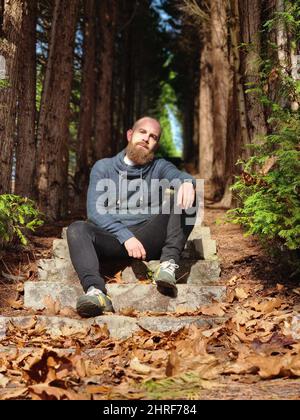Shallow focus of a bearded bald man sitting on park steps lined with trees in autumn Stock Photo