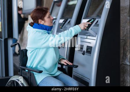 Caucasian woman in a wheelchair buys a train ticket using a mobile phone at a self-service checkout.  Stock Photo