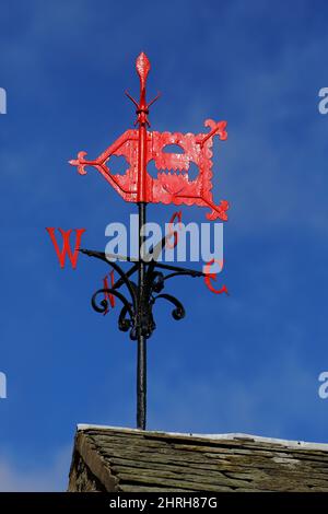 A wind vane, weather vane, or weathercock (black pole with Red flag shaped body and compass point letters) mounted on a slated roof against a blue sky Stock Photo