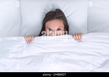 Attractive woman lying on the bed and covering half of face with blanket or hiding under white blanket. Copy space.  Stock Photo