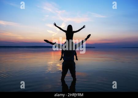 Silhouettes of man and little girl sitting on his shoulders and raising hands Stock Photo