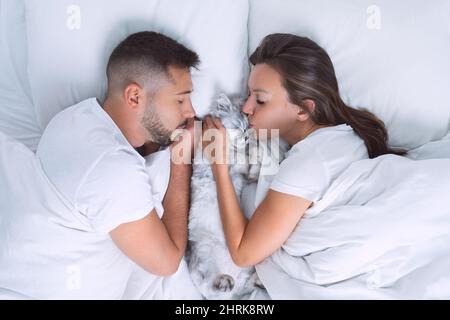 Beautiful couple lying on the bed with their lovely cat. Woman kissing her cute cat. Love for cats or pets. Sleep with cat Stock Photo