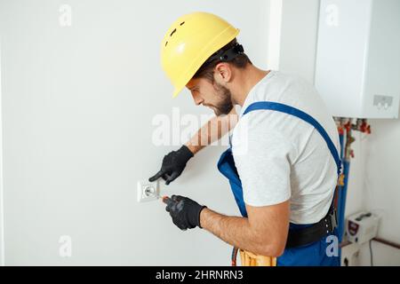 Male worker, professional electrician in hard hat and protective gloves using screwdriver while replacing electrical socket in a room Stock Photo