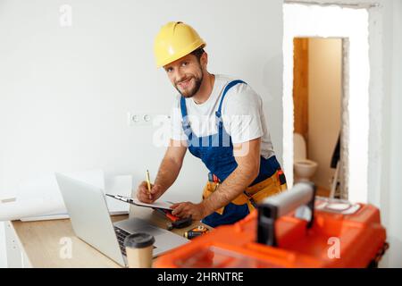 Smiling handyman worker in uniform and hard hat using laptop, writing on a clipboard, looking at camera while standing indoors Stock Photo