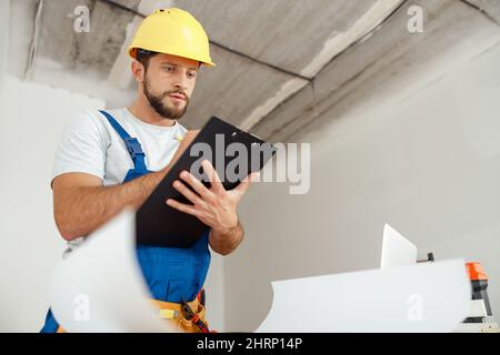 Attentive repairman in workwear and hardhat holding clipboard and making notes while standing indoors during renovation work Stock Photo