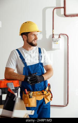 Smiling technician, handyman in uniform, hard hat and protective gloves looking away while standing indoors, leaning on a toolbox Stock Photo