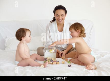 Teaching them responsibility from a young age. A young mother sitting in bed with her two babies busy putting wooden blocks in a jar. Stock Photo