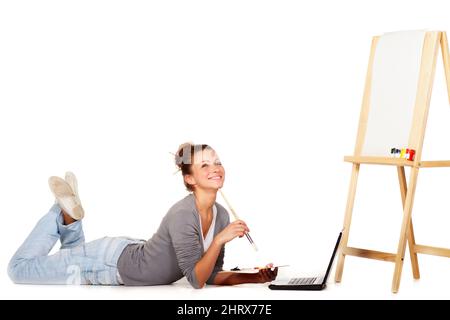 Im looking everywhere for inspiration. Shot of smiling young woman lying down by her laptop and a blank canvas on an easel. Stock Photo