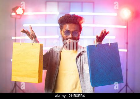 Happy stylish young man on stage showing shopping bags by looking at camera on neaon background - concept of clothing advertisement, promotion offers Stock Photo