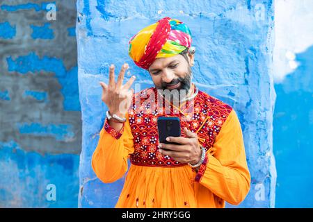 Happy traditional Indian mature man wearing colorful outfits and turban using smart phone, Smiling male holding android smart mobile phone while stand Stock Photo