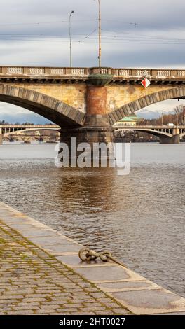 An old mooring ring is fixed on the stone quay on the Vltava river. The pile of the stone bridge is yellow and the two arches start on each side. Stock Photo