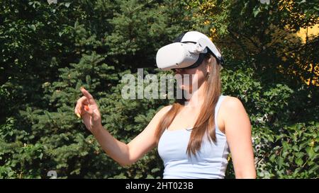 Female in virtual reality glasses controls the device with his hand against the background of green trees Stock Photo