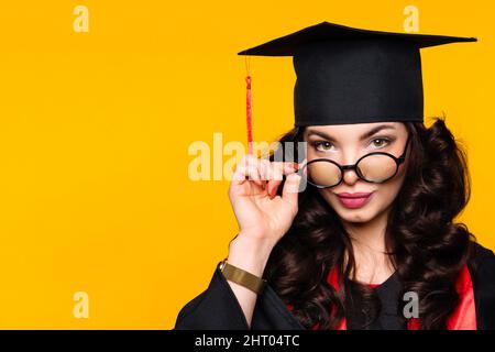 Close up self-confident portrait young woman graduate in graduation hat and eyewear on yellow wall. Female best student wearing graduation cap and ceremony robe holding glasses and looks at camera. Stock Photo