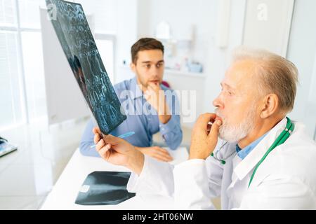 Focused mature adult male physician consult frustrated young man patient giving bad news explaining results of MRI image. Stock Photo