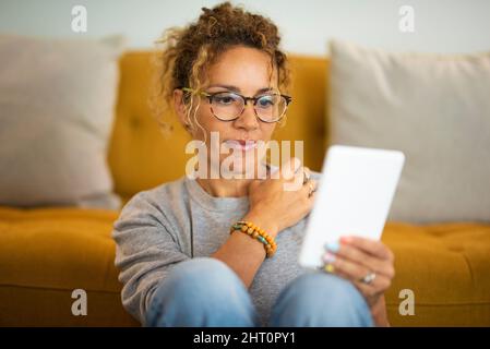 Happy relaxed adult cute woman use electronic device reader to read an e-book and enjoy her indoor leisure activity at home. Pretty female people wear Stock Photo