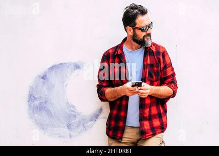 Handsome young adult bearded man use mobile phone connection outdoor sending messages or chatting against a white wall in background. Hipster cute mal Stock Photo