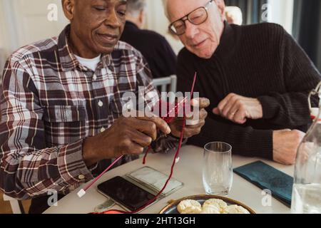 Elderly man knitting by male friend at retirement home