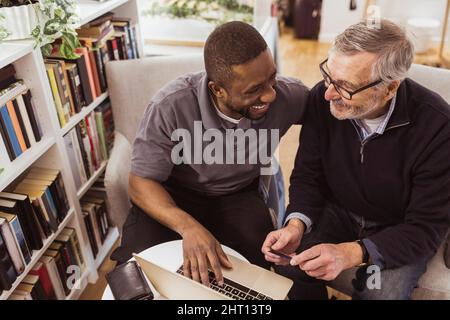 Happy male caregiver teaching online shopping to senior man on laptop at nursing home Stock Photo